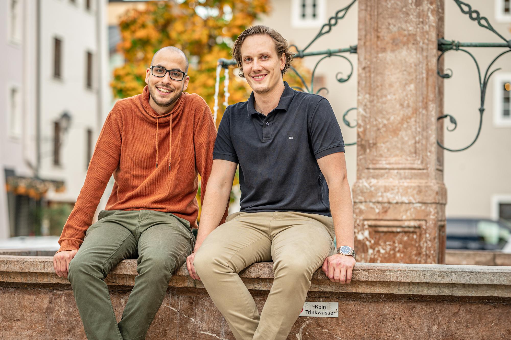 Thomas Steiner und Tobias Franek sitzen auf einem Brunnen mit Blick in die Kamera