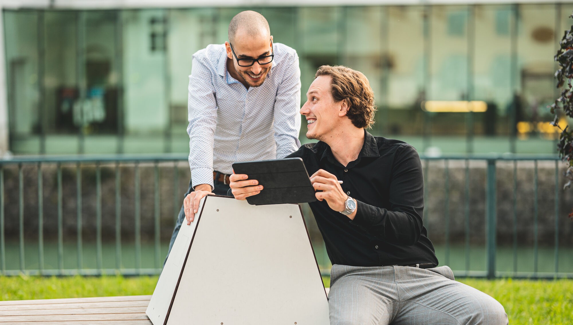 Thomas Steiner sitz auf einer Bank und zeigt Tobias Franek etwas auf einem iPad. Beide lachen.